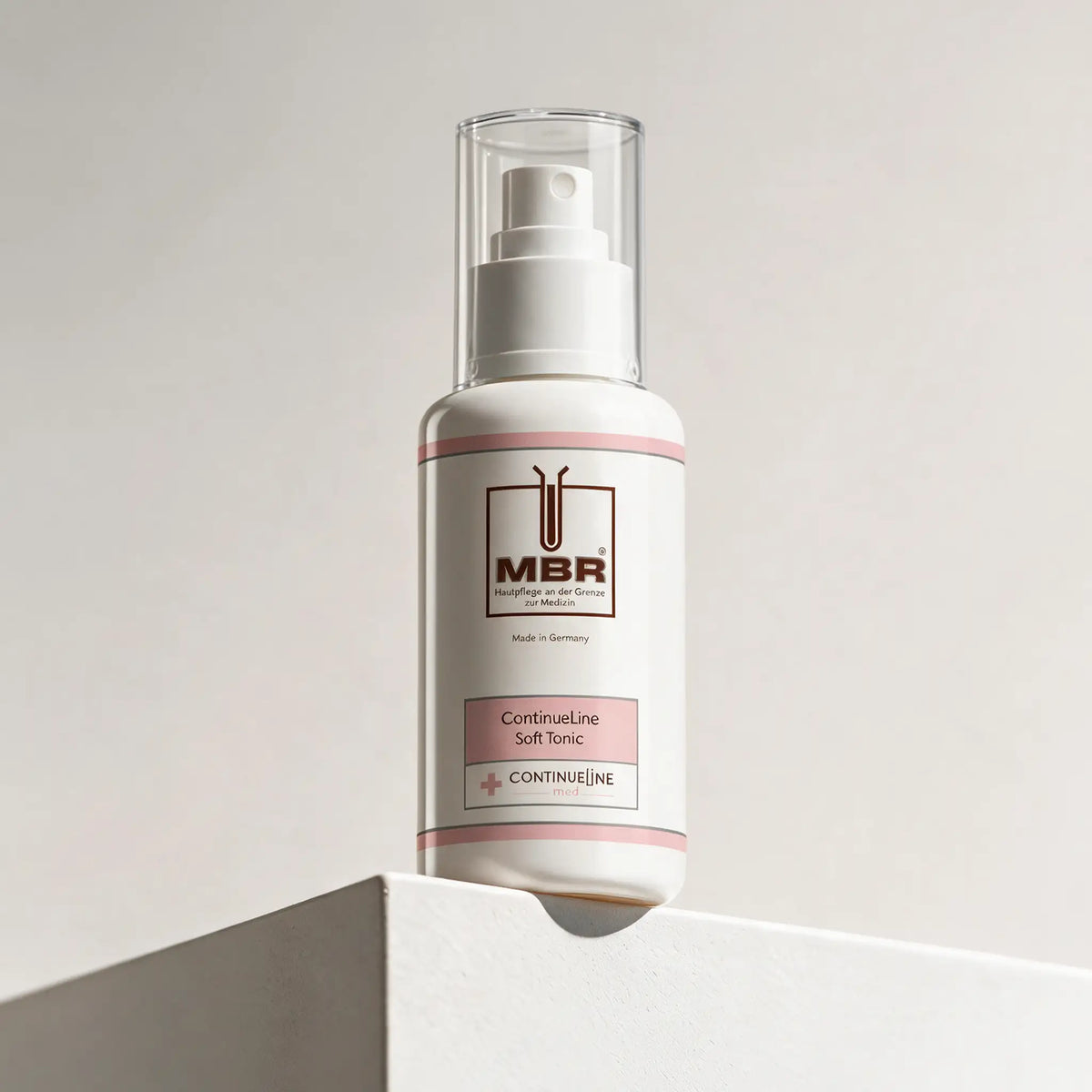 A white pump bottle of MBR Continuline Soft Tonic with pink accents, displayed on a white geometric pedestal against a light beige background.