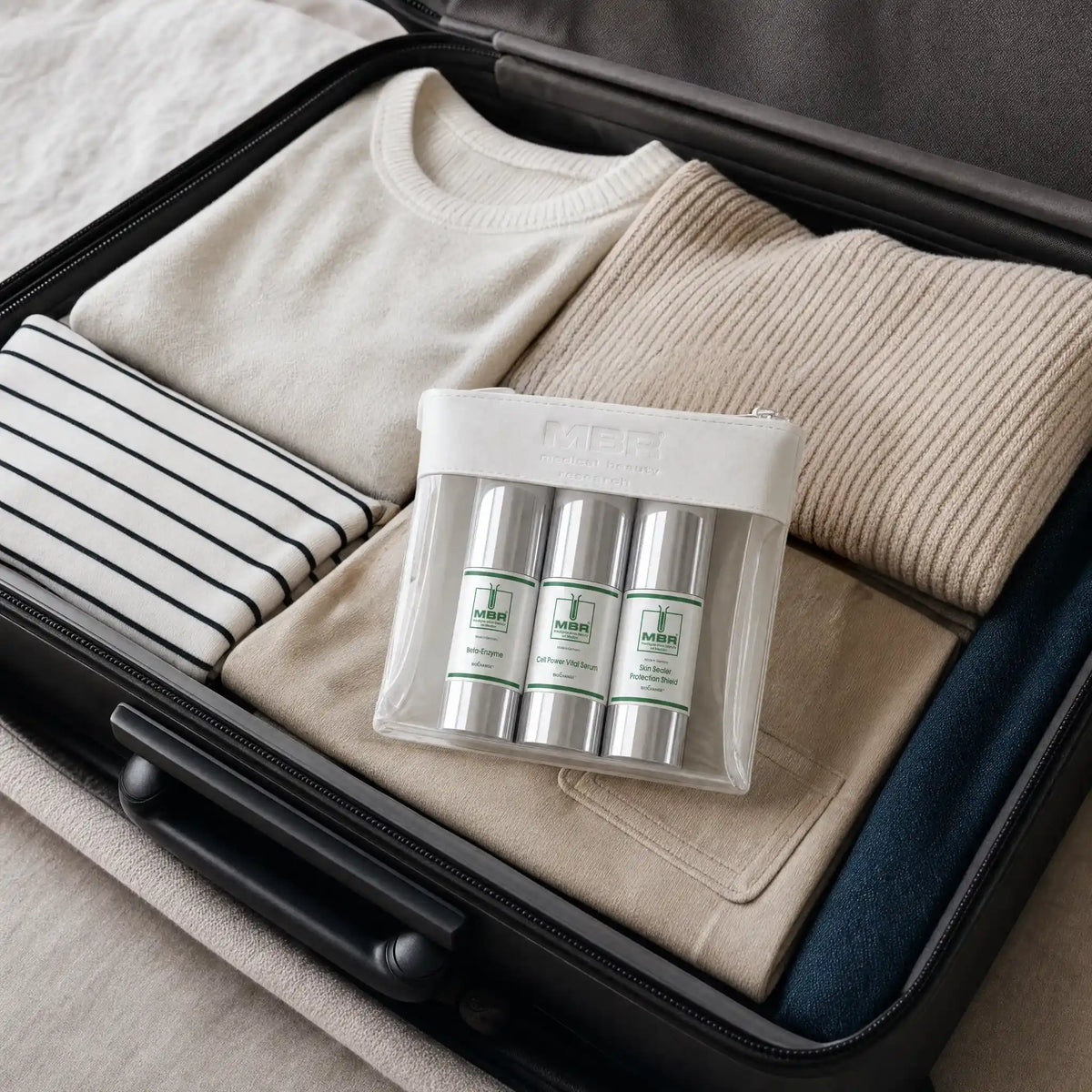 A clear plastic travel pouch containing three silver cylindrical bottles with white and green labels, placed inside an open black suitcase alongside folded beige and navy clothing.