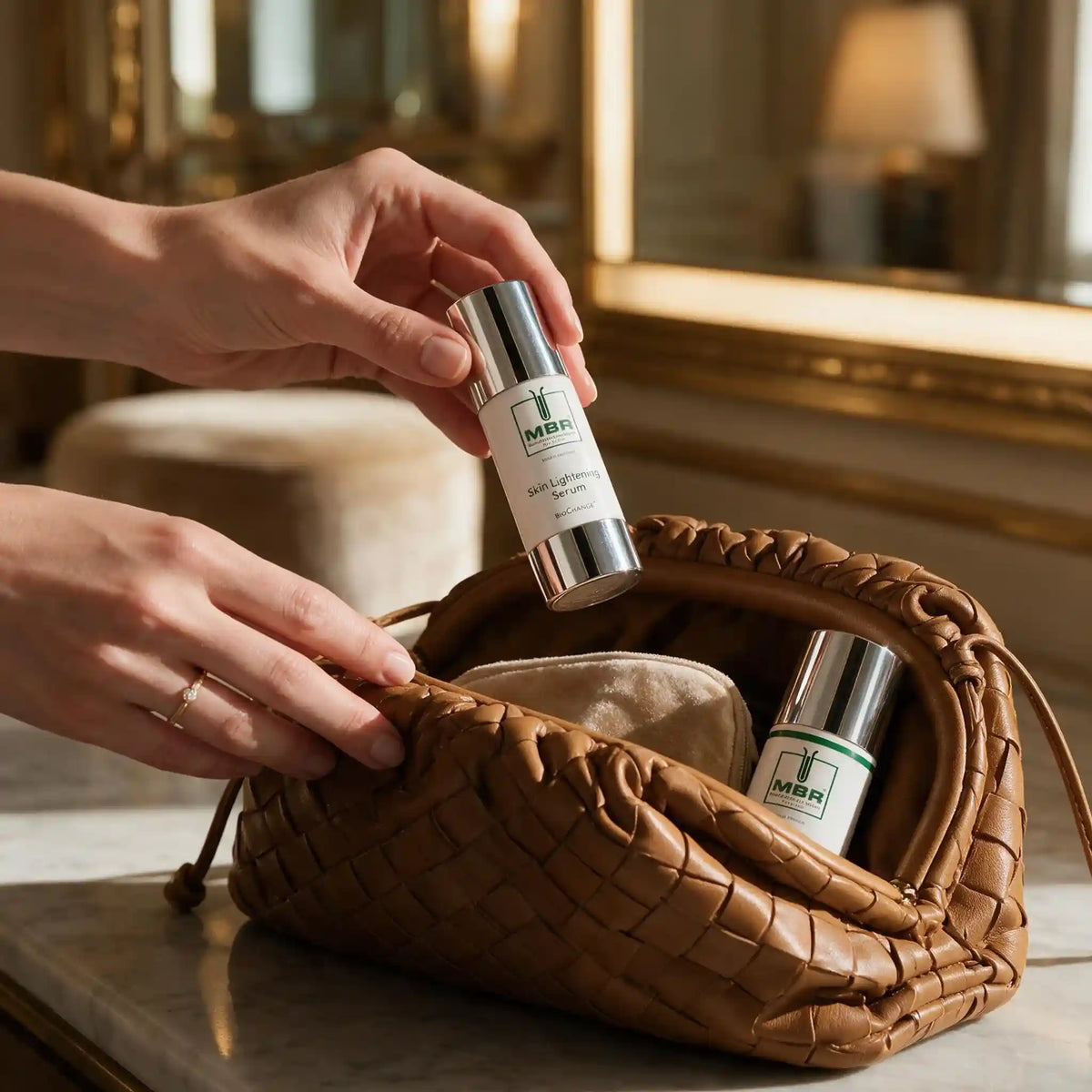 A person's hands are shown holding a small white and silver bottle labeled 'MBR' over an open woven brown leather cosmetic pouch containing two matching bottles.