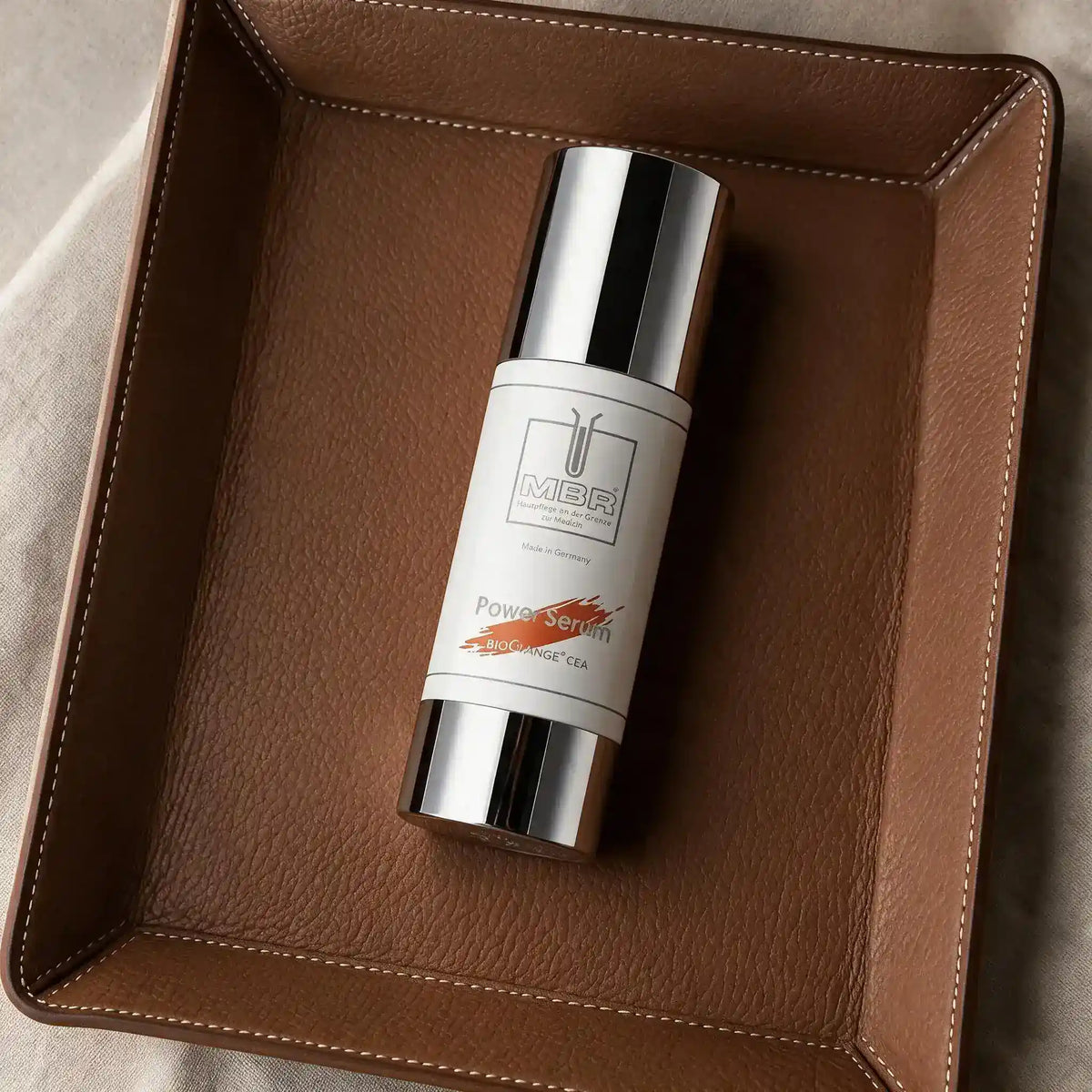 A cylindrical cosmetic bottle with a reflective silver cap and base and a white label reading 'MBR' and 'Power Serum', placed diagonally inside a brown leather tray. The tray has a debossed Hermes Paris logo near the bottom.