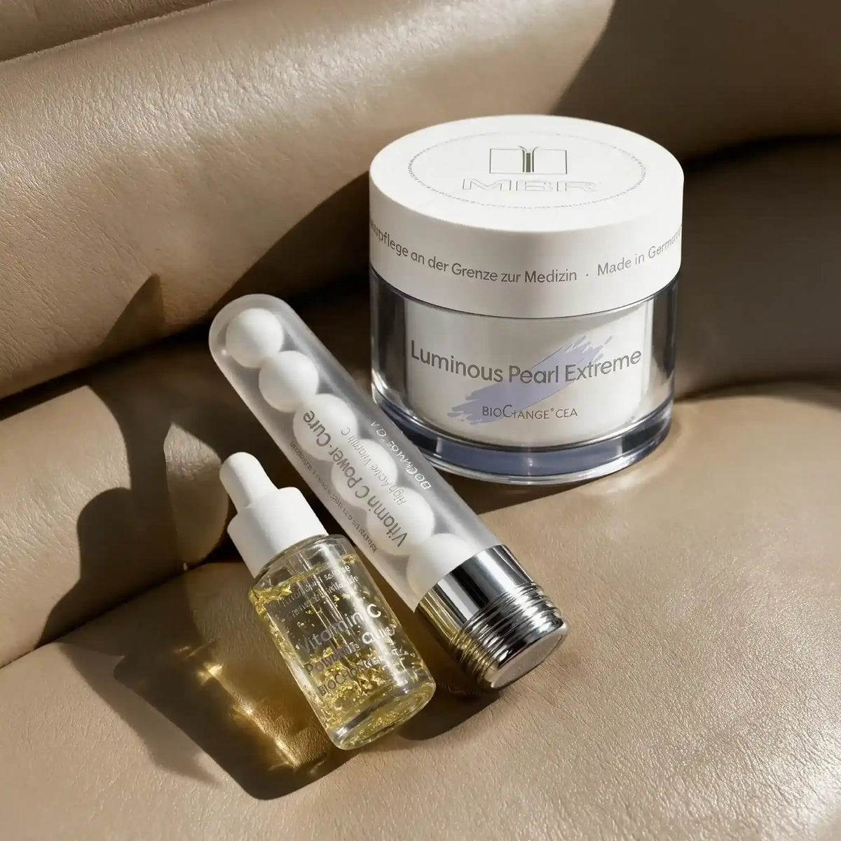 Three skincare products arranged on a beige leather surface: a white jar labeled 'Luminous Pearl Extreme' by MBR, a clear tube labeled 'Vitamin C Bright Care' with white spheres inside, and a small dropper bottle with gold glitter liquid. The scene is styled with warm sunlight and shadows.