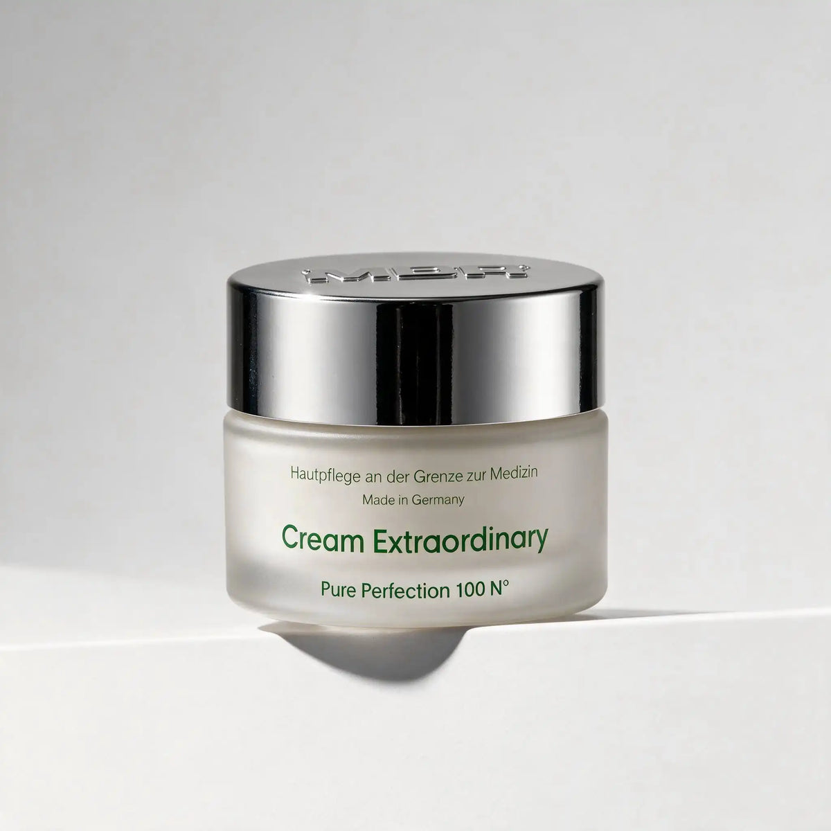 A jar of MBR Extraordinary Pure Perfection 100 N on a clean white surface with a soft shadow. The jar is frosted with green text, topped with a reflective silver cap, and set against a neutral light gray background.