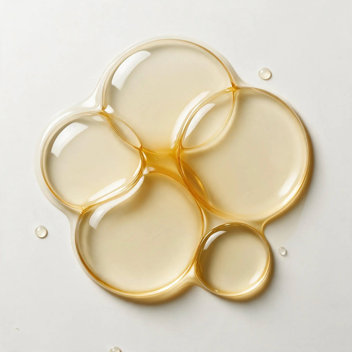 A glossy, translucent pool of pale golden liquid, resembling honey, oil, or gel, arranged in organic circular shapes on a clean white background.