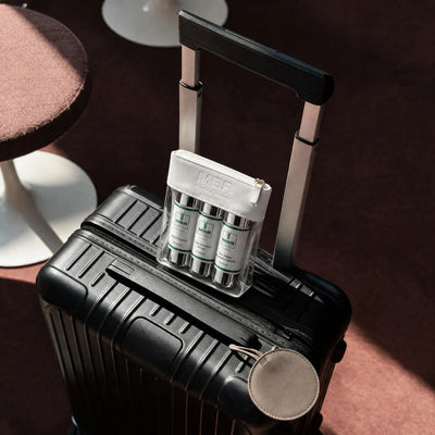 A black ribbed hard-shell suitcase with an extended handle sits on a warm brown surface. On top of the suitcase is a clear organizer holding three white skincare bottles with green MBR labels, and a small round beige pouch hangs from a strap.