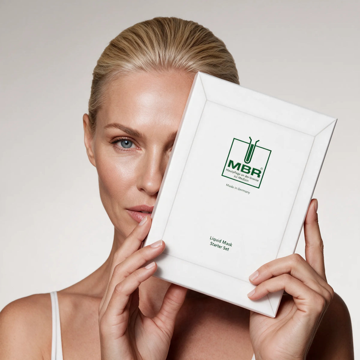A blonde woman holds a white MBR Liquid Mask Staple Set box in front of her face against a light gray studio background. [AI]