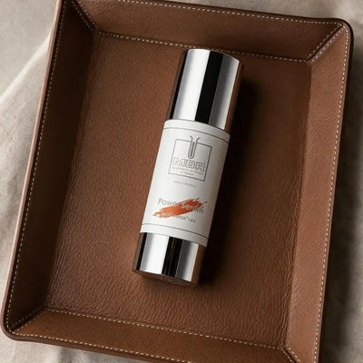 A cylindrical cosmetic bottle with a reflective silver cap and base and a white label reading 'MBR' and 'Power Serum', placed diagonally inside a brown leather tray. The tray has a debossed Hermes Paris logo near the bottom.