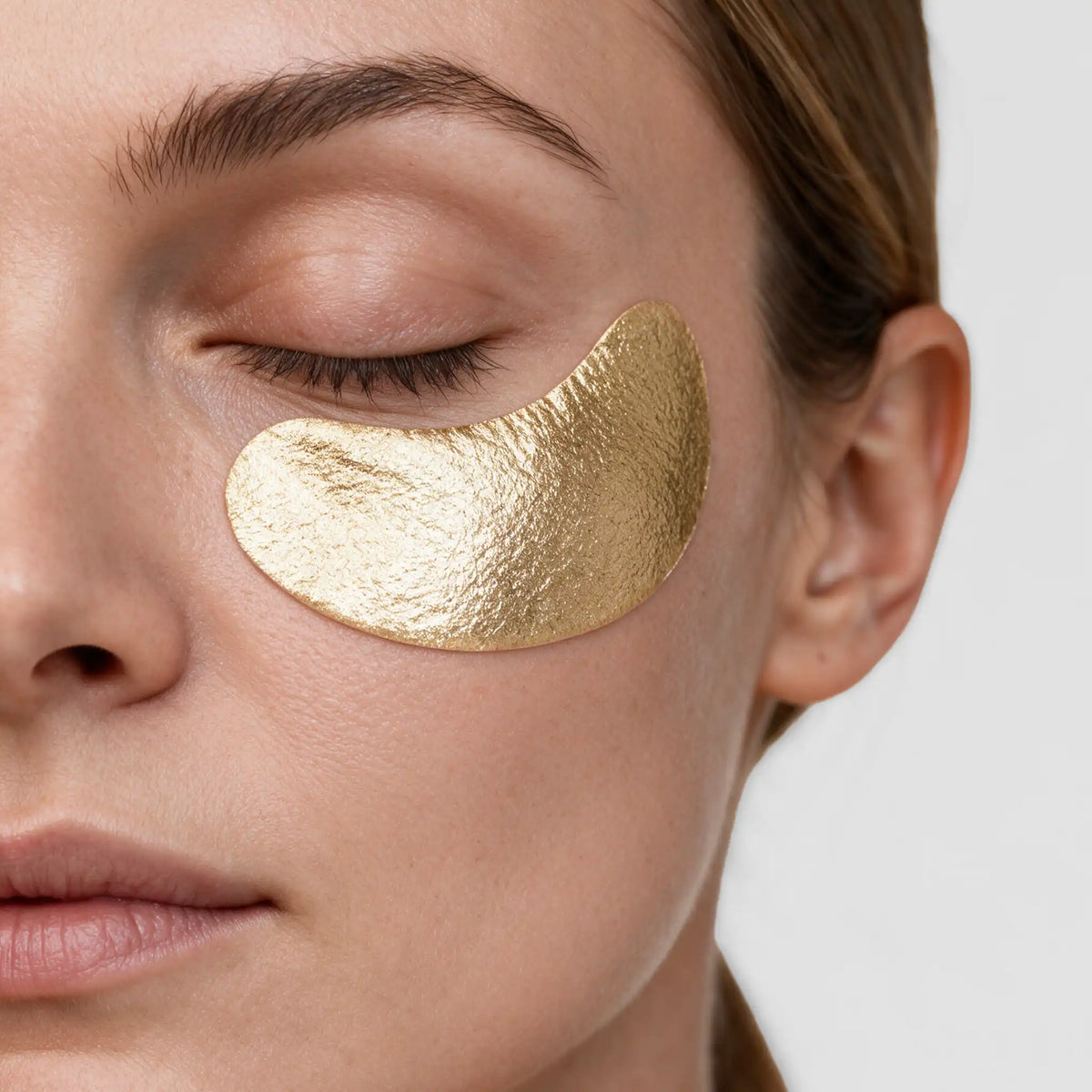 Close-up of a woman's face with a metallic gold eye patch applied under her closed eye, shown against a clean light gray background. [AI]