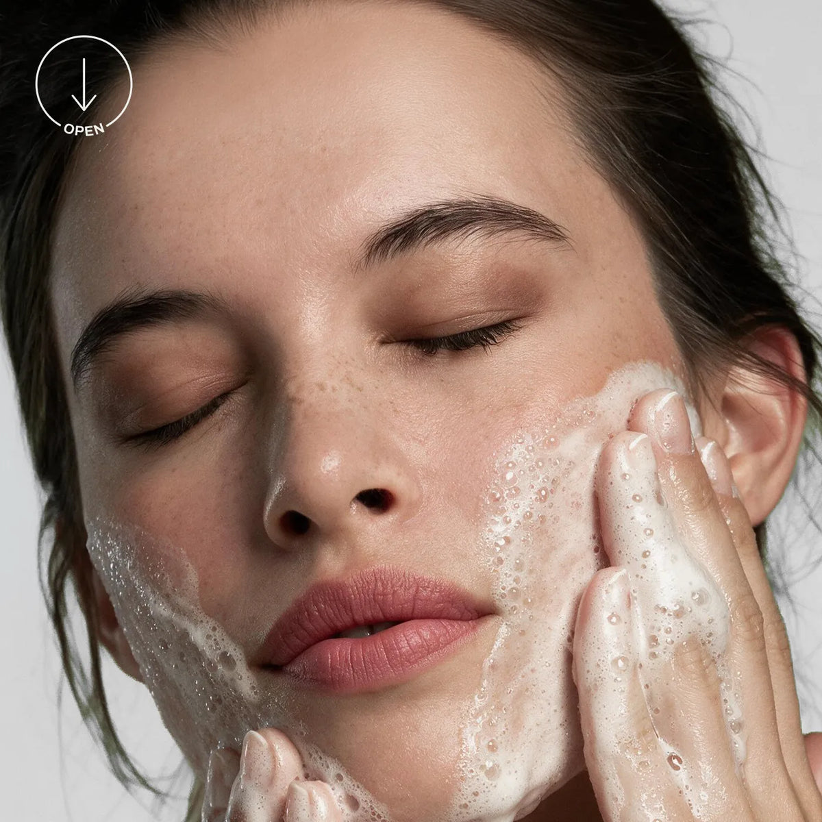 Close-up of a woman with closed eyes applying or washing foam onto her face, with her hands touching her cheek. A small 'OPEN' icon appears in the top-left corner. [AI]
