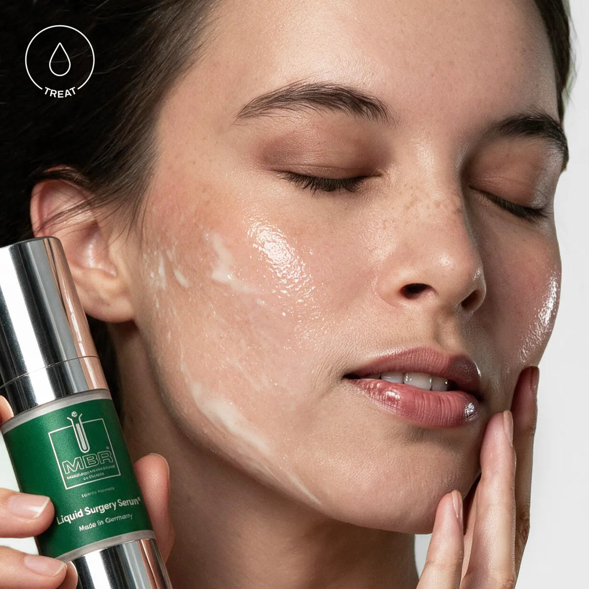 A close-up of a woman with a glossy, hydrated complexion applying or absorbing a white cream on her cheek, while holding a green and silver bottle of MBR Liquid Surgery Serum. [AI]